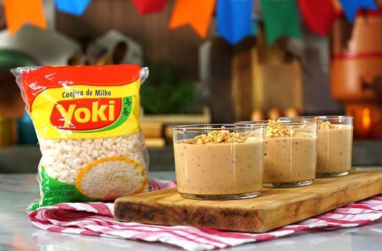 Potinhos de canjica de doce de leite sobre uma tábua ao lado de pacote de canjica de milho Yoki, em cenário de festa junina