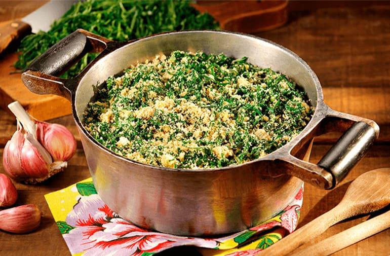 Farofa de couve mineira em panela de ferro, acompanhada de couve fresca e cabeças de alho na mesa