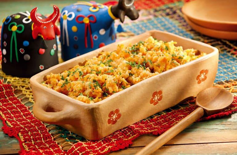 Farofa de jerimum em travessa com desenho de flores, em mesa com bois decorativos coloridos