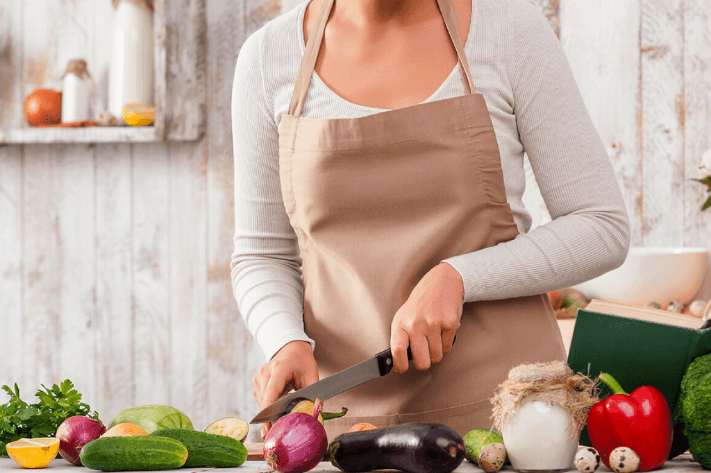 Uma pessoa na cozinha, usando um avental e cortando uma cebola. Vários vegetais, como alface, tomates, pepinos e uma berinjela, estão espalhados na bancada.