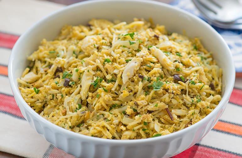 Bacalhau a Bras em uma travessa de vidro branca, com batata palha, azeitonas, salsa e pedacinhos de cebola e alho