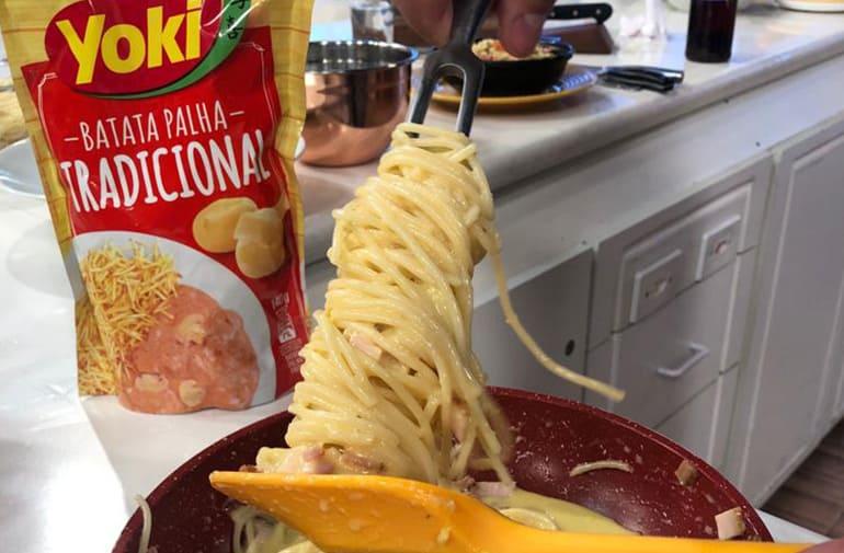 Macarrão carbonara cremoso servido direto da panela, ao lado de embalagem de batata palha Yoki Tradicional