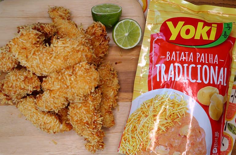 Frango empanado crocante ao lado de pacote de batata palha Yoki, servido em tábua de madeira com limão cortado