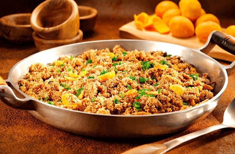 Panela de ferro com farofa de frango com pequi, uma colher de ferro, cumbucas de madeira e pequis sobre uma tábua ao fundo