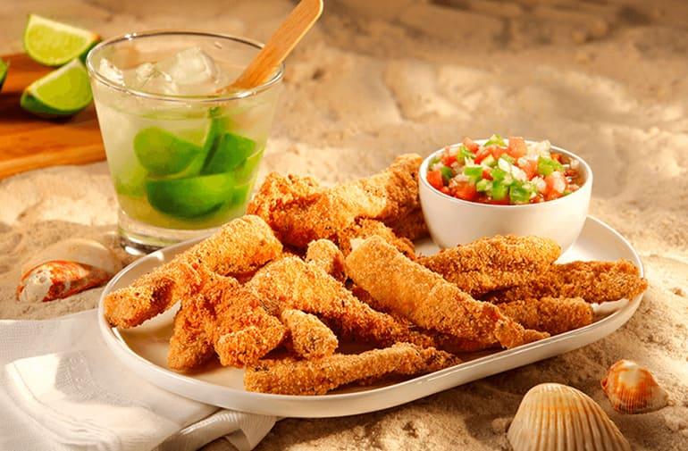 Aperitivo de peixe empanado com farofa Yoki, acompanhado de vinagrete, na areia da praia, com uma caipirinha de limão