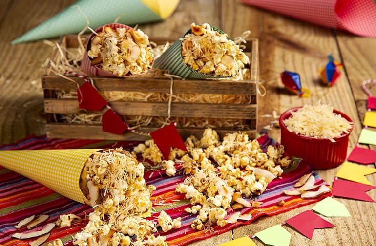 Pipoca queijadinha com amêndoas, servida em cones coloridos, em uma mesa com caixa rústica e decoração de festa junina