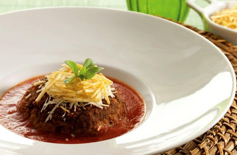 Polpettone de carne com molho sugo, coberto com parmesão e batata palha, em prato fundo de porcelana