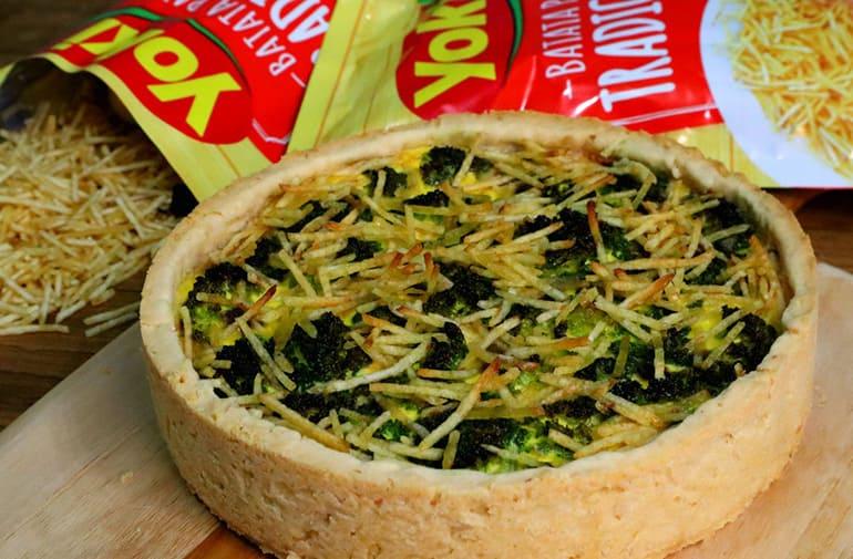 Quiche de brocolis com recheio crocante, servido em tábua de madeira ao lado de pacotes de batata palha Yoki