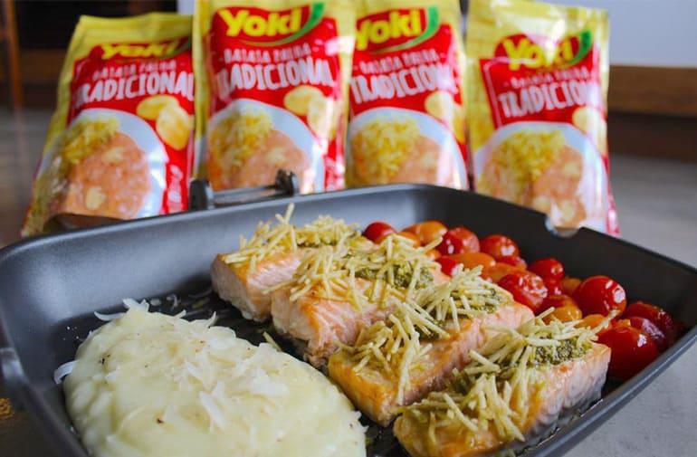 Salmão grelhado com batata palha, tomate confit e purê de batatas cremoso, servidos em forma ao lado de pacotes de batata palha Yoki