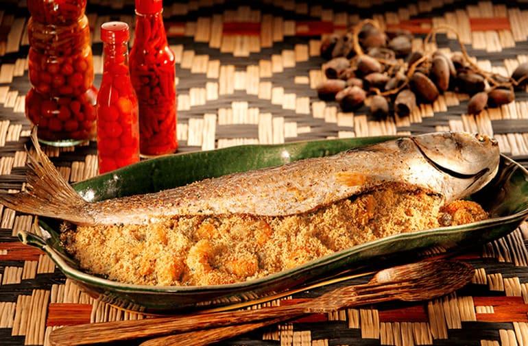 Tainha frita sobre farofa de camarão, em prato com formato de folha de bananeira, mesa tropical com potes de pimenta