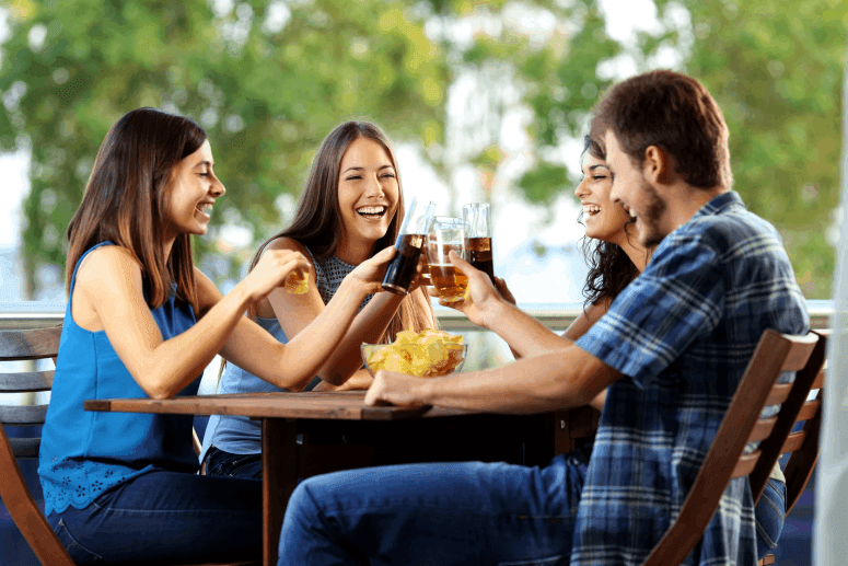Quatro amigos alegres brindam em uma mesa de bar, com um recipiente de petiscos recheado com Yokitos no centro